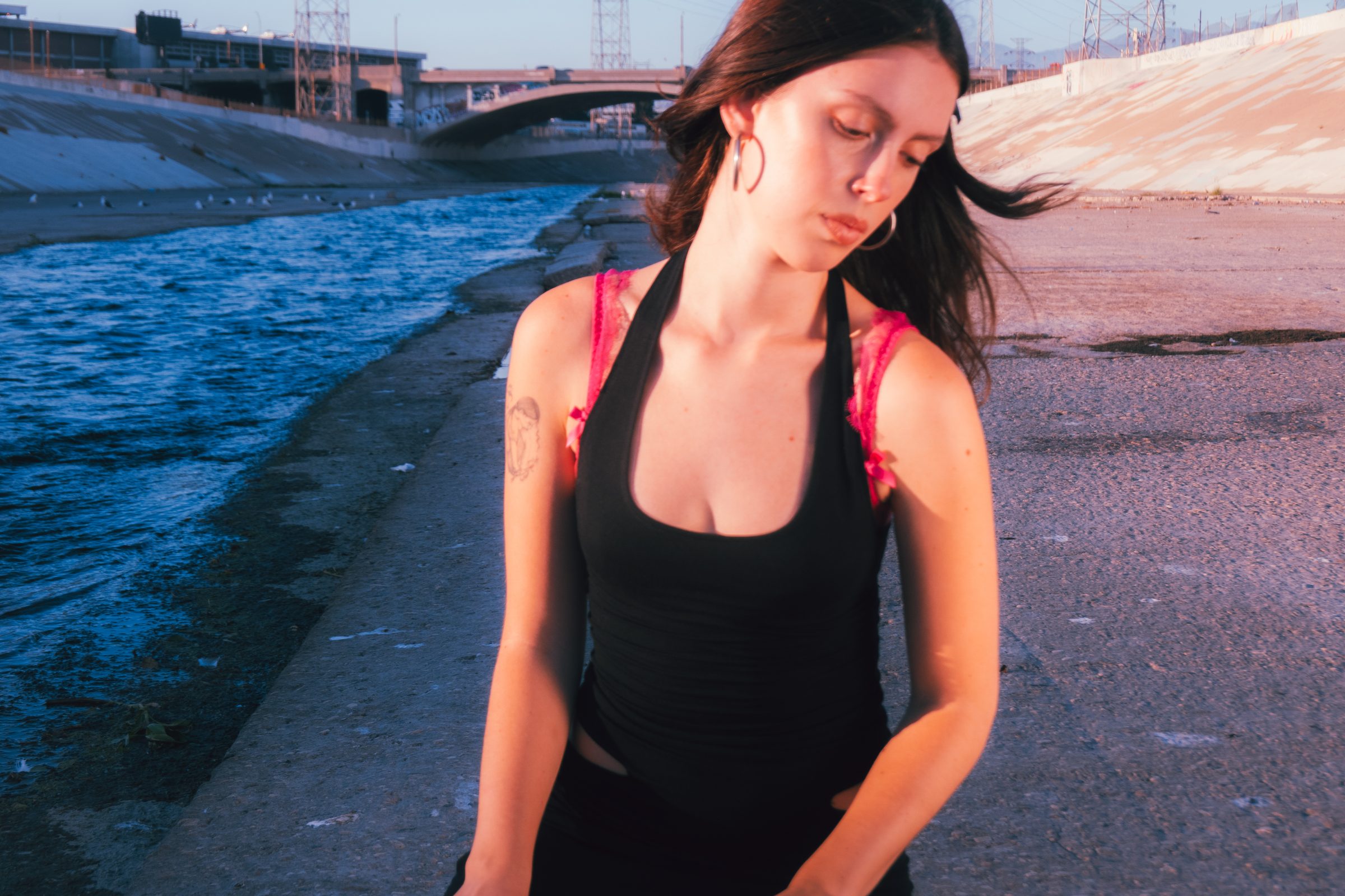 Melina Suriani standing along the LA river at golden hour, wearing a black tank top with pink lace straps.