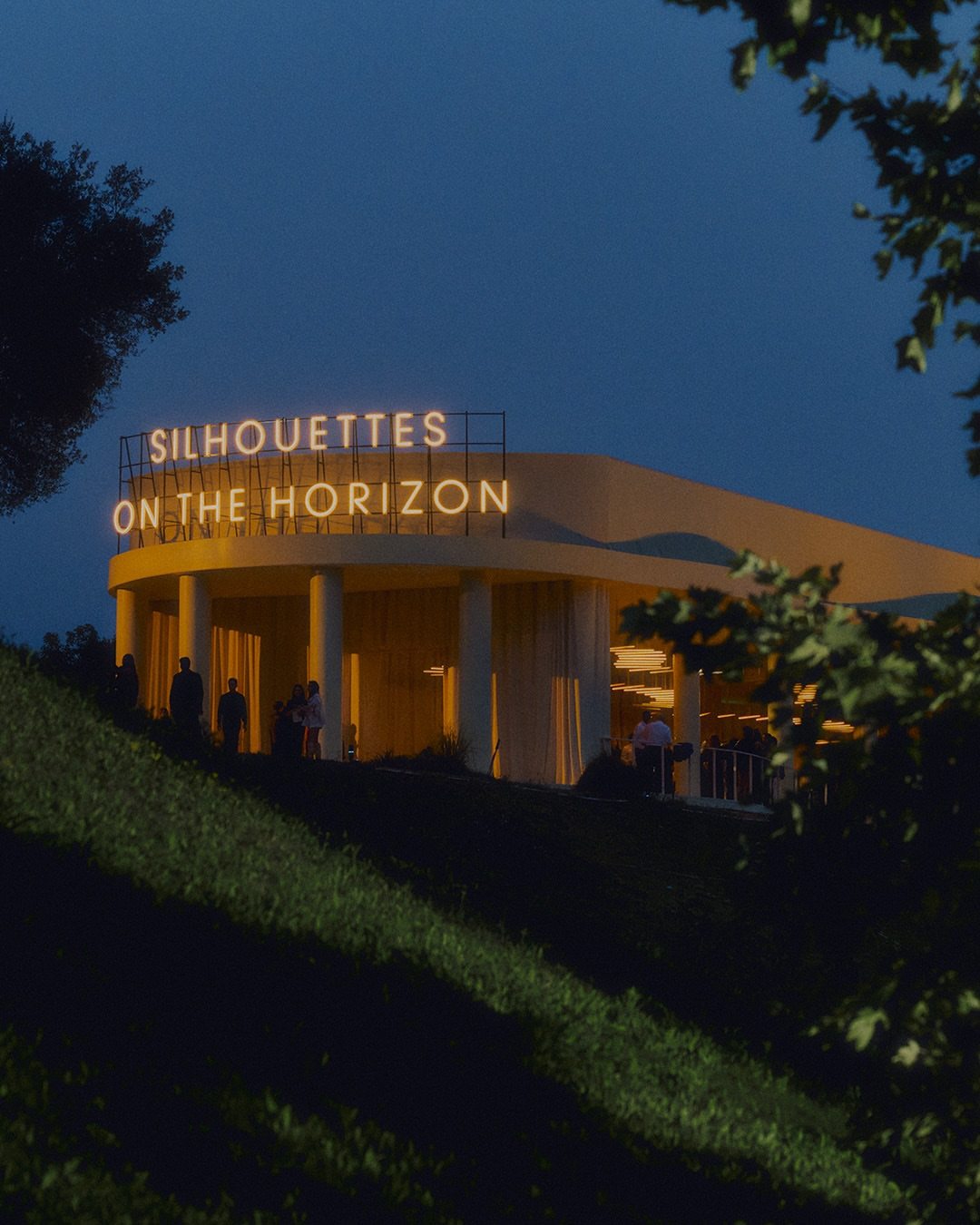 Hermès show venue at dusk, “Silhouettes on the Horizon” lit sign above a colonnaded pavilion on a hillside