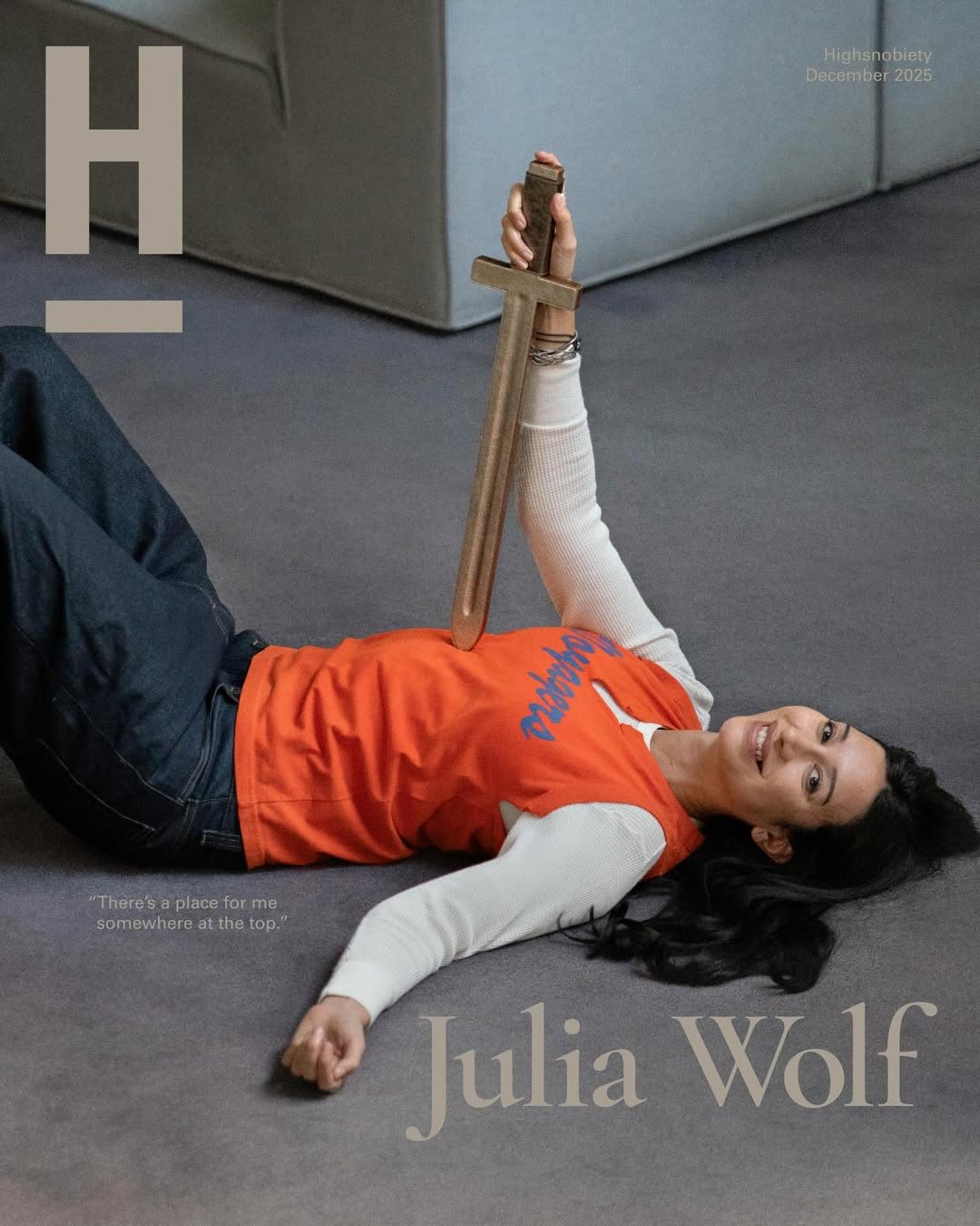 Julia Wolf x Highsnobiety cover, lying on a grey carpet holding a wooden sword upright, orange tee over a white thermal