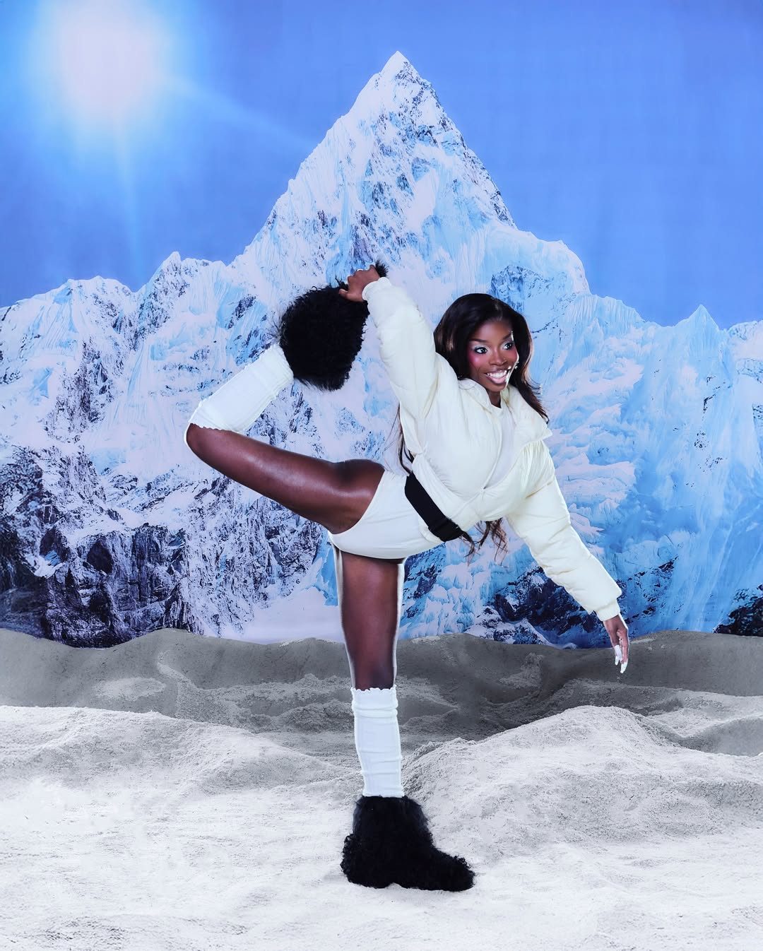 Model in a cream cropped puffer and black shaggy boots holding a standing leg stretch in front of the mountain backdrop